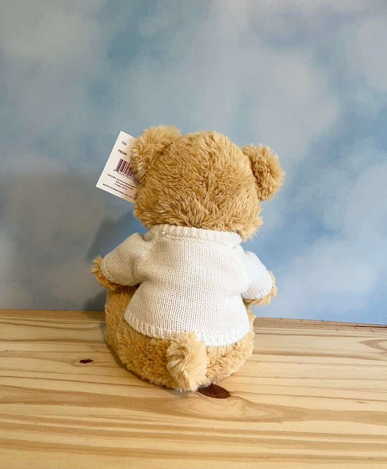 May include: A plush teddy bear, viewed from the back, wearing a white sweater. The bear has light brown fur and is sitting on a wooden surface. A tag is attached to the bear's ear. The background is a light blue sky.