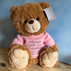 Könnte beinhalten: Ein brauner Plüschteddybär, der einen hellrosa Strickpullover trägt. Der Pullover ist mit dem Text Always with us Grandma bestickt, gefolgt von den Daten 07-07-1929 und 07-27-2003. Der Bär hat ein weißes Etikett an seinem Ohr befestigt.