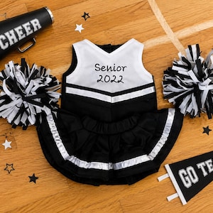 May include: A black and white cheerleader outfit featuring a sleeveless top with the text Senior 2022, a pleated skirt, two pom-poms, a black megaphone with the word CHEER, and a black pennant flag with the text GO TEAM! and white star accents.