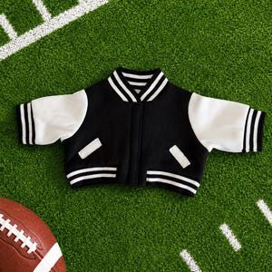 May include: A black and white varsity jacket featuring a black body, white sleeves, and a ribbed collar, cuffs, and hem with white stripes. The jacket includes two white-trimmed pockets on the front and a central snap-button closure system.