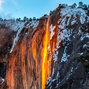 May include: A fiery orange waterfall cascades down a snow-capped cliff face, creating a striking contrast against the gray rock. The waterfall appears to be illuminated from behind, creating a glowing effect.
