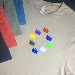 Può includere: Una maglietta beige con un design di sette corone colorate in cerchio. Le corone sono di colore rosso, viola, giallo, nero, blu, bianco e verde.