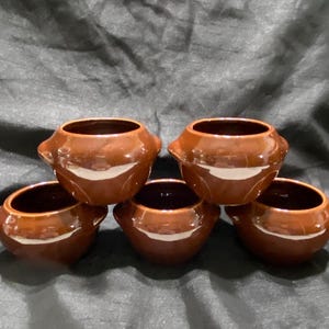 以下が含まれることがあります： 5つの小さな茶色のセラミックボウルのコレクション。各ボウルは丸みを帯びたボディ、暗い内側、2つの小さなハンドルを備えています。ボウルは積み重ねられた形で配置されており、上に2つ、中央に2つ、下に1つあります。