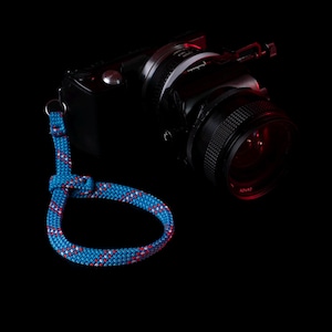 Könnte beinhalten: Eine schwarze Kamera mit einer blau-rot gemusterten Handschlaufe. Der Gurt besteht aus einem gewebten Seilmaterial. Das Kameraobjektiv ist groß und schwarz.