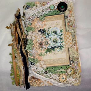 Könnte beinhalten: Ein handgefertigtes Journal mit floralem Design. Die Hülle zeigt eine Collage aus Spitze, Stoff und Verzierungen in Beige-, Grün- und Cremetönen. Eine Quaste hängt an der Seite. Das Journal ist mit Knöpfen und einem Blumendruck verziert.