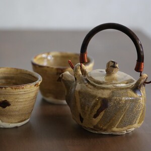 Può includere: Un set da tè in ceramica con una teiera con manico scuro e due tazze abbinate. Il set ha un'estetica rustica e terrosa con toni marroni e beige. La teiera ha un coperchio e un beccuccio. Le tazze sono piccole e cilindriche.