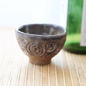 Op de afbeelding: Een klein, bruin keramiek sake-kopje met een swirl-patroon. Het kopje staat op een bamboemat.