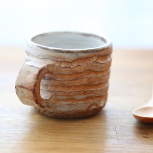 Op de afbeelding: Een witte keramische mok met een bruine, getextureerde buitenkant. De mok heeft een afgeronde handgreep en staat op een houten oppervlak.