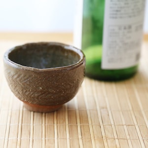 Op de afbeelding: Een kleine, bruine keramische kom met een getextureerd patroon staat op een geweven bamboemat. De kom is leeg en lijkt van aardewerk te zijn.