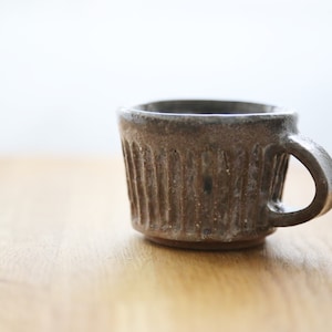 Op de afbeelding: Een kleine, grijze keramische mok met een getextureerd, geribbeld ontwerp en een handvat. De mok staat op een houten oppervlak.