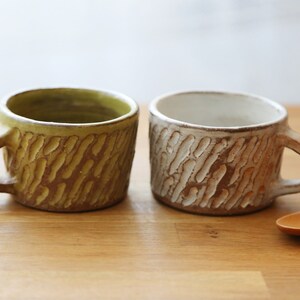 Op de afbeelding: Twee keramische mokken met een getextureerd, golvend ontwerp. De ene mok is groen en de andere is wit. De mokken staan op een houten oppervlak.