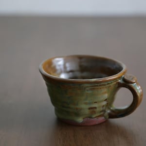 Könnte beinhalten: Handgefertigte Keramiktasse mit einzigartigem, strukturiertem Design. Die Tasse hat eine grüne und braune Glasur mit einem kleinen Henkel. Der Boden hat einen rosafarbenen Farbton. Ein rustikales, handwerkliches Stück.