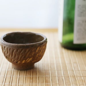 Op de afbeelding: Een klein, bruin, keramisch sake-kopje met een getextureerd oppervlak. Het kopje staat op een bamboemat.
