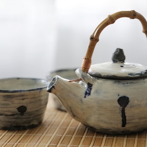Può includere: Un set da tè in ceramica con una teiera con manico in bambù e due tazze abbinate. La teiera e le tazze sono bianco sporco con dettagli blu e neri. Il set è appoggiato su un tappetino di bambù.