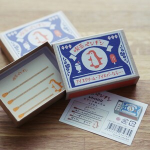 Pode incluir: Uma caixa de fósforos azul e branca com uma ilustração de pinguim e o texto "喫茶ペンギン" (Kissa Penguin) e "アイスクリーム・アイスバーなら…" (Sorvete e picolés se…). A caixa de fósforos está aberta e mostra um pequeno cartão com uma ilustração de pinguim e o texto "レトロ日記" (Diário retro).