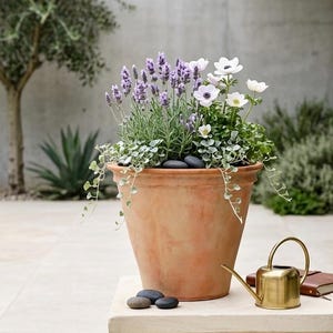 May include: A terracotta plant pot overflowing with lavender, white flowers, and greenery, placed on a stone plinth. A small brass watering can and a brown notebook are beside the pot. The scene is set outdoors with a tree and other plants in the background.