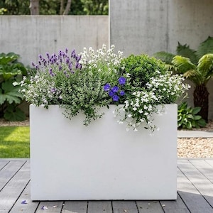 May include: A white rectangular planter box overflowing with a mix of flowering plants. The plants feature purple lavender, white blooms, and blue flowers, with green foliage. The planter sits on a wooden deck, with a concrete wall and greenery behind.