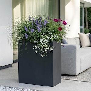 May include: A dark grey rectangular planter overflowing with colourful flowers and greenery. The flowers include purple, pink, and white blooms. The planter is on a patio next to a modern outdoor seating area.