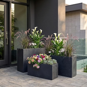 May include: Three dark grey rectangular planters filled with various plants and flowers. The planters are arranged on a paved patio near a glass door. The plants include white calla lilies, ornamental grasses, and colourful petunias.