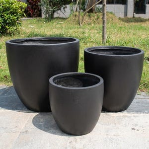 May include: Three black, rounded planters of varying sizes are arranged outdoors. The planters have a matte finish and a simple, modern design. They are set on a stone surface with grass and foliage in the background.