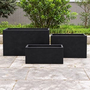 May include: A set of three rectangular planters in a matte black finish. The planters are arranged in a tiered formation, with the largest in the back left, a medium-sized one in the back right, and the smallest one positioned in the front centre.
