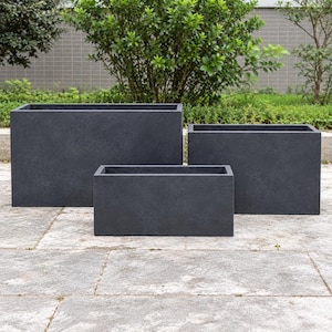 May include: A set of three rectangular planters in a dark charcoal grey finish. The planters are arranged in a tiered formation, featuring smooth, solid walls and open tops, designed for holding plants in an outdoor setting.