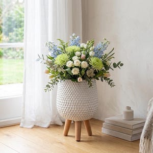May include: A white, textured ceramic vase with a honeycomb design, standing on three wooden legs, displays a colourful floral arrangement. The bouquet includes white roses, green chrysanthemums, blue delphiniums, and foliage. The vase is positioned near a window with white curtains.