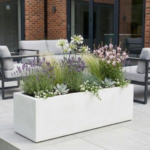 May include: A white rectangular concrete planter filled with a variety of plants and flowers. The planter contains lavender, white, and pink blooms, along with green foliage. Outdoor patio furniture is visible in the background.
