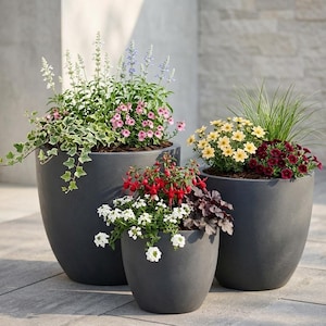 May include: Three dark grey, oval-shaped planters filled with colourful flowers and greenery. The plants include pink, white, yellow, and red blooms, with trailing ivy and tall grasses. The planters are arranged on a stone patio.