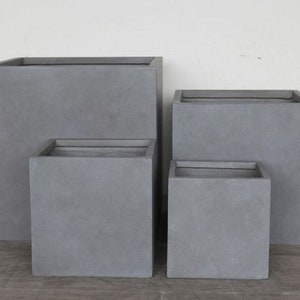 May include: A set of four square planters in varying sizes, each featuring a smooth, matte grey finish. The planters are arranged in a cluster, with two larger units positioned in the back and two smaller units placed in the front, all with open tops.