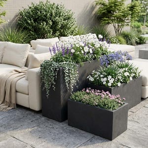 May include: An outdoor patio scene featuring a cream-coloured sectional sofa and several dark grey rectangular planters filled with colourful flowers and greenery. The scene is set on a stone patio with lush landscaping in the background.