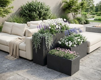 Puede incluir: Escena de patio al aire libre con un sofá seccional color crema y varias jardineras rectangulares de color gris oscuro llenas de flores y vegetación coloridas. La escena está ambientada en un patio de piedra con exuberante paisajismo de fondo.