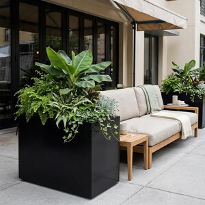 May include: An outdoor patio scene with a black rectangular planter filled with green plants. A light-coloured sofa with a wooden side table and a blanket is to the right. The scene is set on a concrete patio.