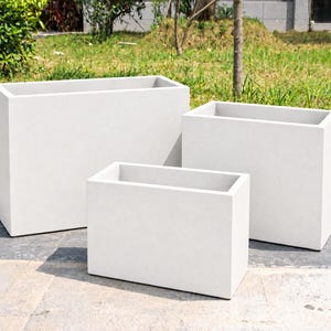 May include: Three rectangular planters in varying sizes, each featuring a smooth, matte white finish. These open-top containers have clean, sharp edges and a solid, uniform construction, arranged in a cluster on a paved surface.