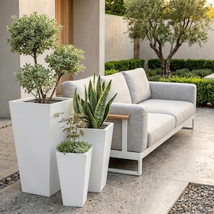 May include: A modern outdoor patio setting with a light grey sofa and white metal frame. Three white square planters contain greenery. The scene is set on a concrete patio with a stone border, creating a minimalist aesthetic.