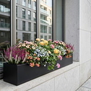 May include: A black rectangular planter box filled with a variety of colourful flowers, including pink, yellow, and purple blooms. The planter sits on a ledge in front of a window, with a building in the background.