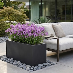 May include: A rectangular black planter box filled with vibrant purple phlox flowers, positioned on a patio surface surrounded by smooth grey river stones, adjacent to a light grey outdoor sofa with matching throw pillows and a small side table.