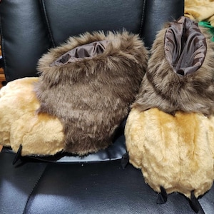 May include: A pair of plush, novelty slippers designed to resemble animal paws. The slippers feature a light brown paw with black claws and a dark brown, furry upper section. The interior lining is a dark brown satin-like material.