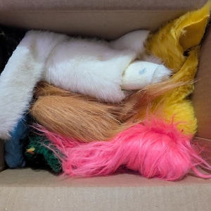 May include: A cardboard box containing assorted faux fur pieces in a variety of colours. The fur includes white, pink, yellow, and brown. The pieces are likely for a costume or craft project.
