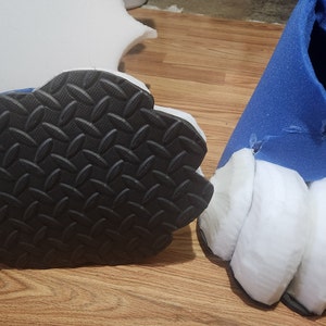 May include: Close-up of a pair of furry blue and white feet with black paw pads. The feet are made of foam and are likely part of a costume.