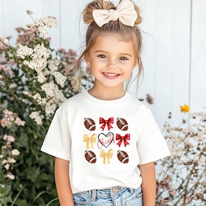 May include: A white t-shirt with a red heart and the word "Chiefs" inside. The shirt is decorated with yellow and red bows and brown footballs.