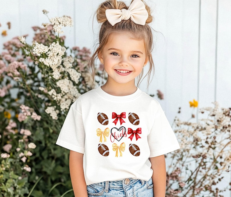 May include: A white t-shirt with a red heart and the word "Chiefs" inside. The shirt is decorated with yellow and red bows and brown footballs.