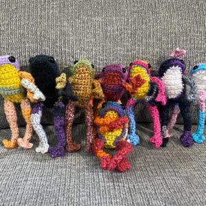 May include: A group of ten crocheted frog plushies in various colours, including purple, yellow, green, red, orange, and blue. The frogs are all standing on a grey sofa.