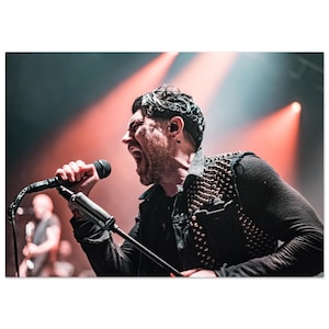 May include: A man with a microphone in hand sings into the microphone while performing on stage. He is wearing a black shirt and a vest with studs. The stage is lit with red and white spotlights.