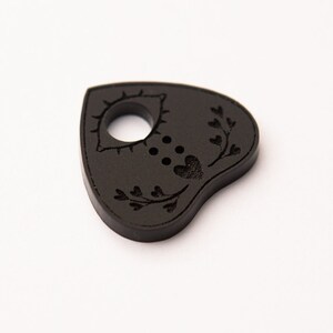 Könnte beinhalten: Schwarze Holzplanchette mit einem Herz- und Augenmotiv. Die Planchette ist herzförmig und hat ein Loch in der Mitte.