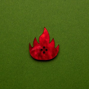 May include: A red wooden button shaped like a flame with four holes in the center. The button is on a green background.