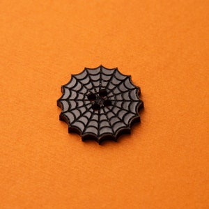 May include: Black spiderweb button with a raised design. The button is on a solid orange background.