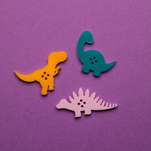 Peut inclure: Trois boutons en forme de dinosaures de couleurs jaune, turquoise et rose sur fond violet. Chaque bouton comporte quatre petits trous. Les dinosaures sont un Tyrannosaurus Rex, un Brontosaurus et un Stégosaure.