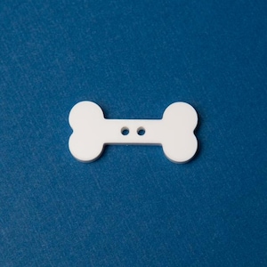 May include: White bone-shaped button with two holes in the center. The button is on a blue background.