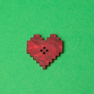 Pode incluir: Um botão vermelho em forma de coração com quatro furos no centro.